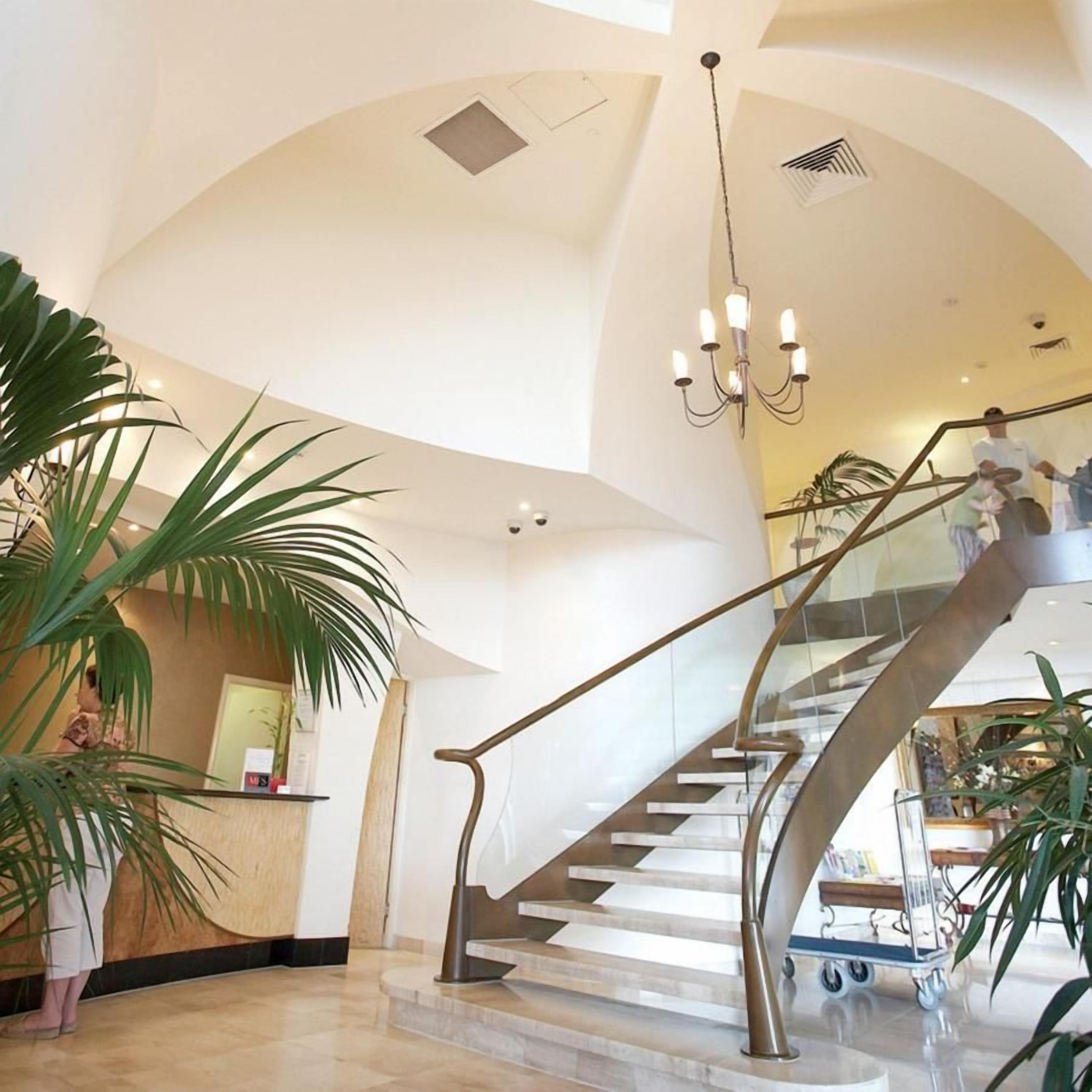 A spacious lobby with a grand staircase, a reception desk, and large potted plants.
