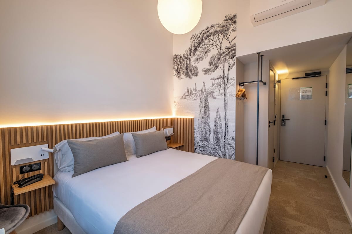 A modern hotel room with a large bed, wooden headboard, bedside table with a lamp, and a wall mural.