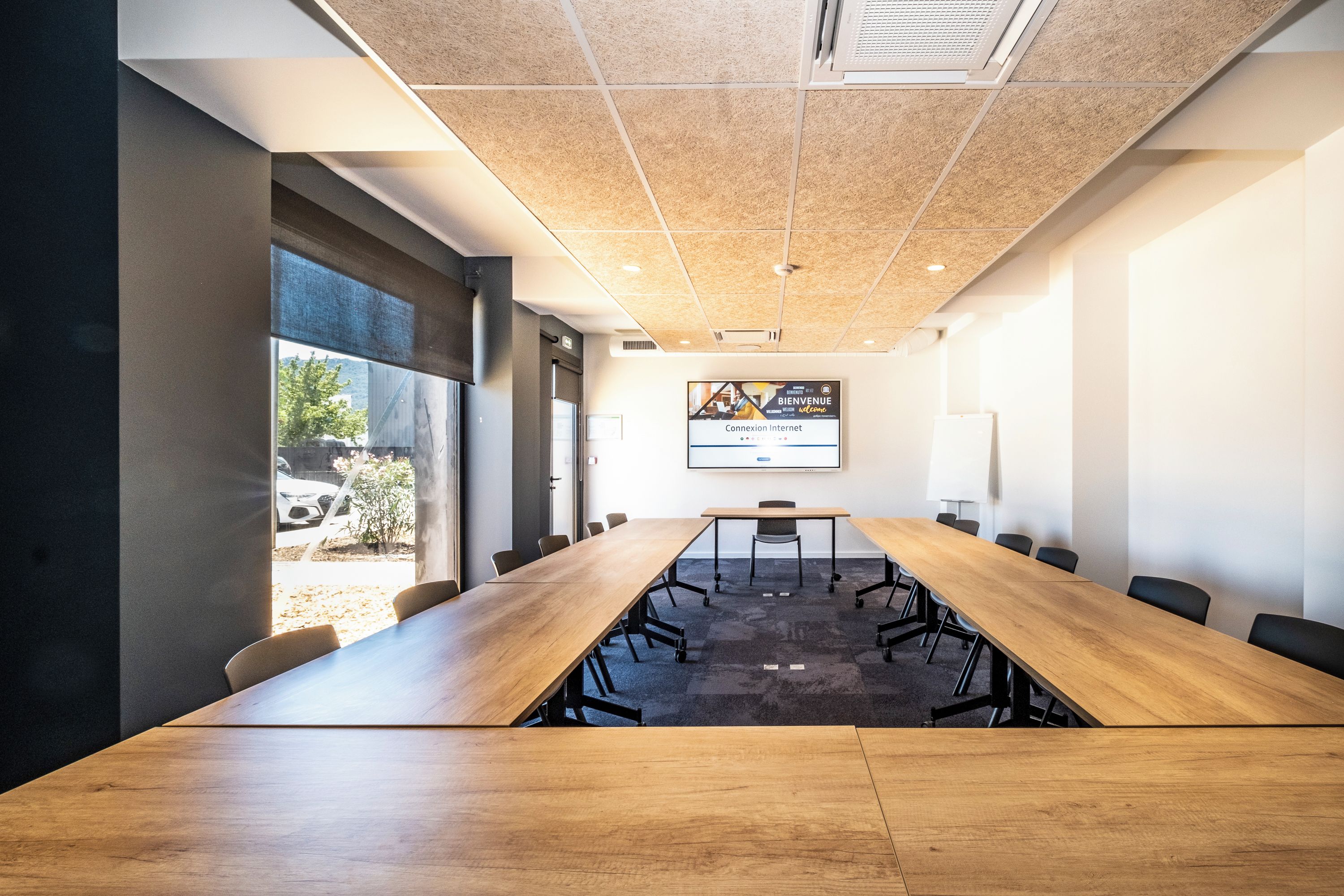 Une salle de conférence avec une longue table en bois, des chaises noires et un écran de projection affichant un message de bienvenue.