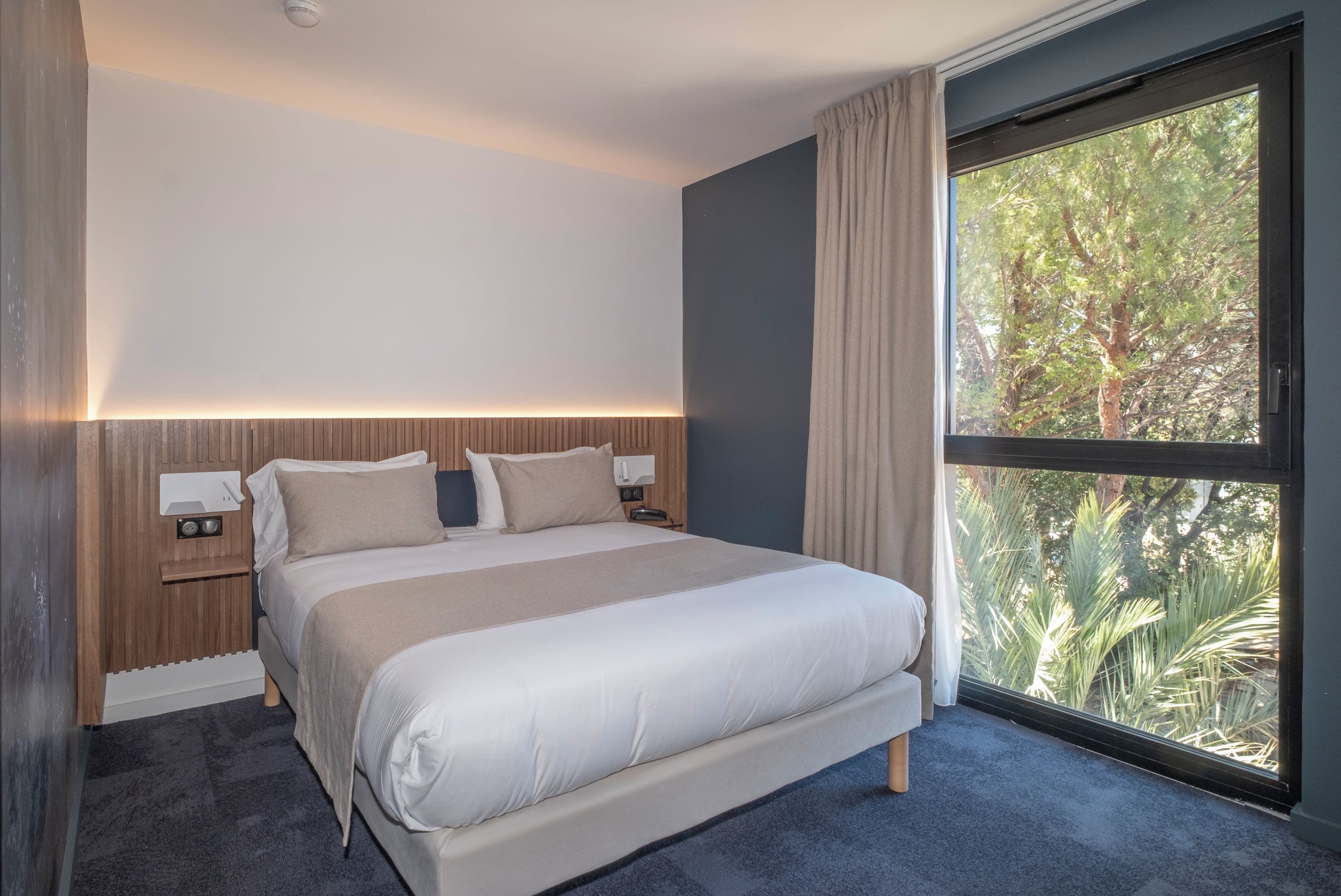 Habitación de hotel moderna con una cama grande, una mesita de noche con lámpara y una ventana con vistas a árboles.