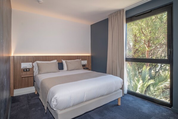 Habitación de hotel moderna con una cama grande, una mesita de noche con lámpara y una ventana con vistas a árboles.