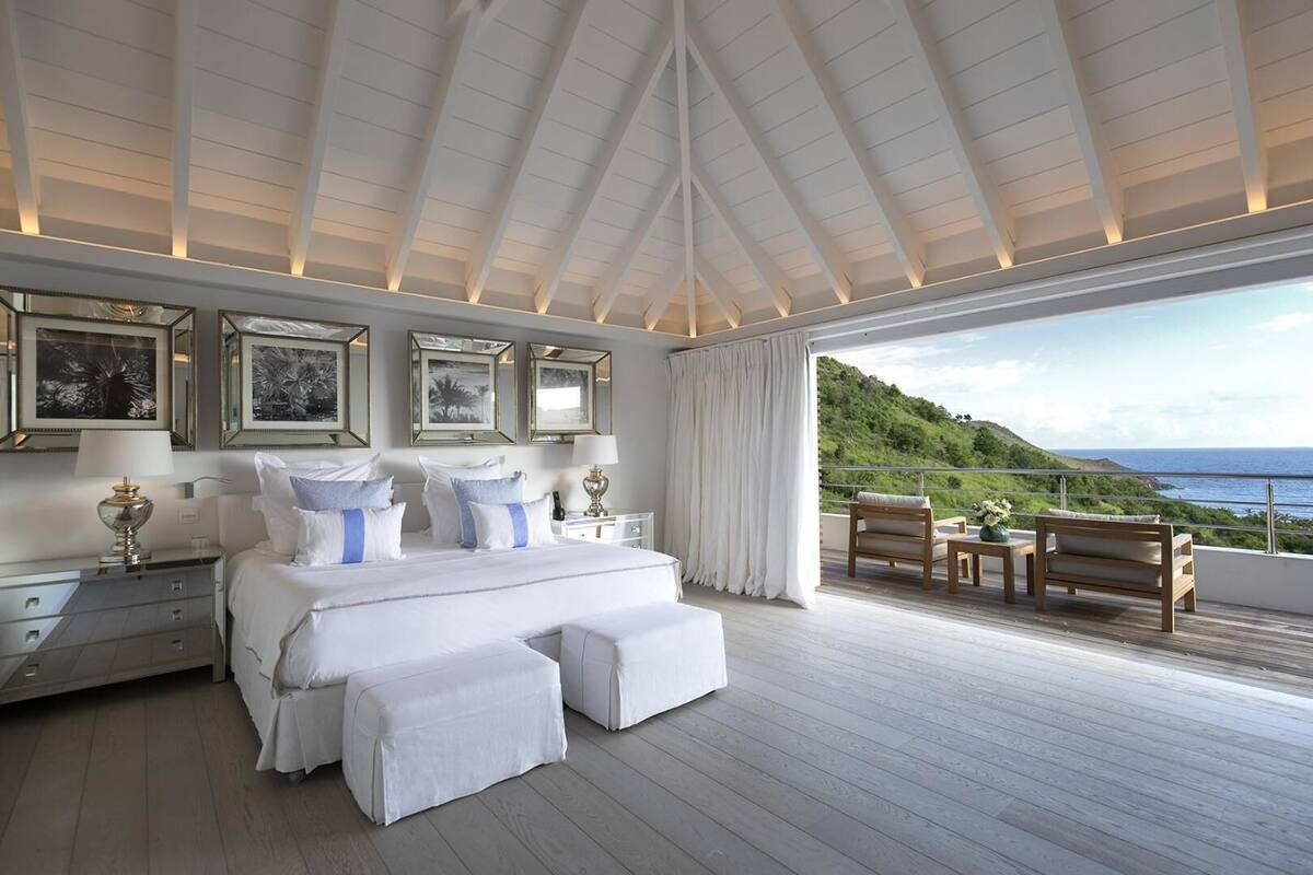 A spacious bedroom with a large bed, a balcony with wooden furniture, and framed artwork on the wall.