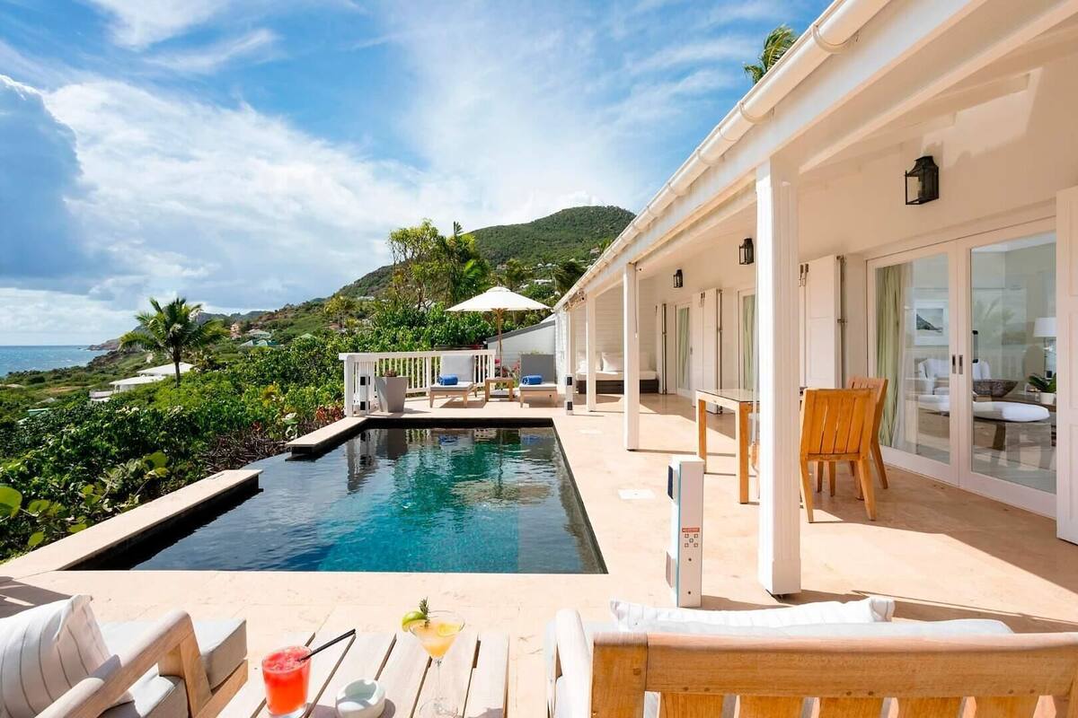 A poolside area with lounge chairs, a table with drinks, and a view of the ocean and hills.