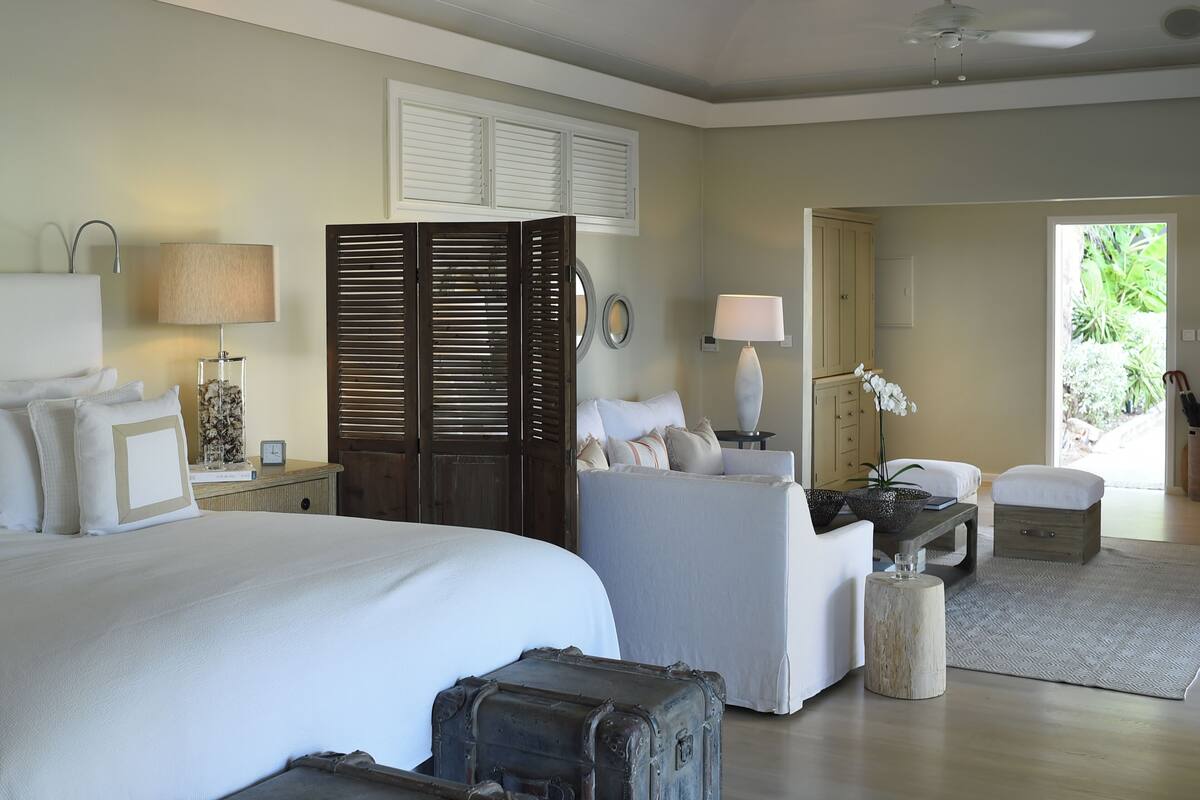 A hotel room with a bed, a sofa, a desk, a lamp, a window with shutters, and a view of greenery outside.