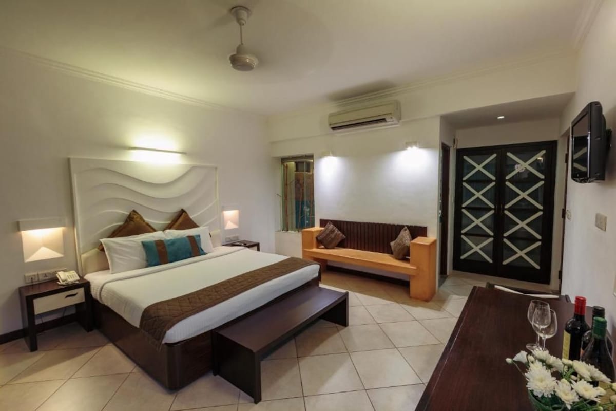 A hotel room with a bed, bedside tables, a bench, a sofa, a TV, and a door with a window.