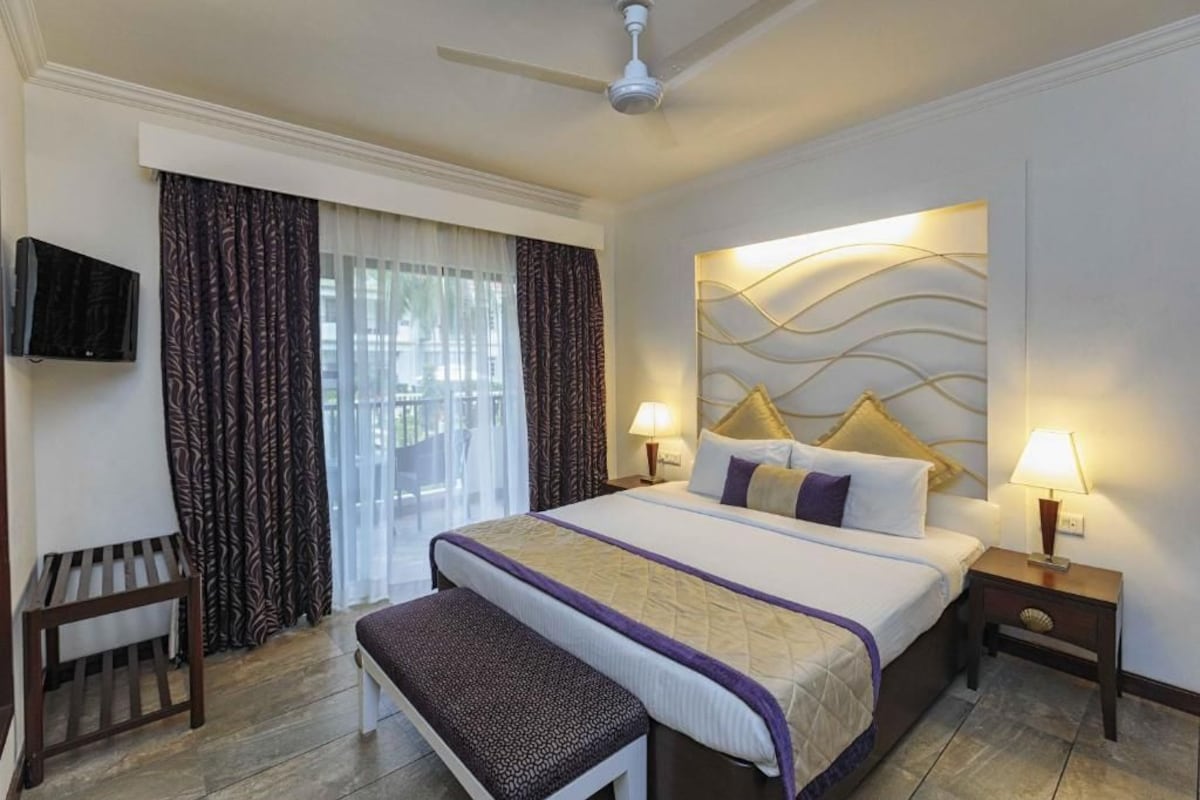A hotel room with a bed, two bedside lamps, a television, a bench, and a ceiling fan.