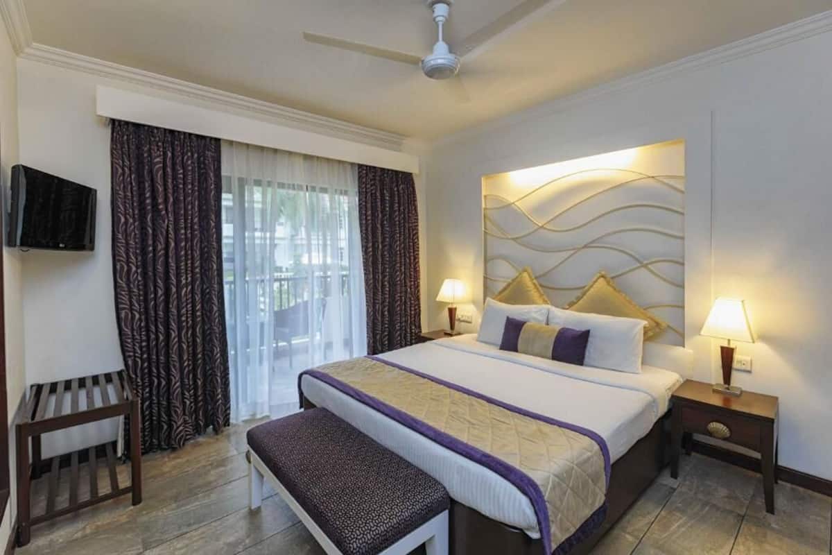 A hotel room with a bed, two bedside lamps, a television, a bench, and a ceiling fan.