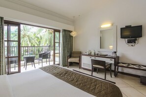 A hotel room with a bed, a desk, a chair, a television, and a balcony with chairs and curtains. - Sonesta Inns Beach Resort - Candolim Beach (Candolim)