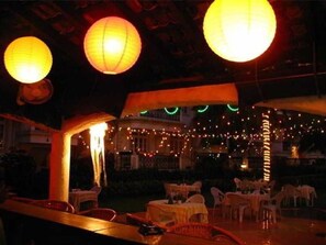 Un espacio exterior para comer con mesas y sillas, iluminado por faroles colgantes y luces de cadena.