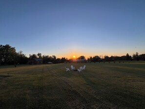 A grassy field with a sunset in the background, a few chairs, and a building.