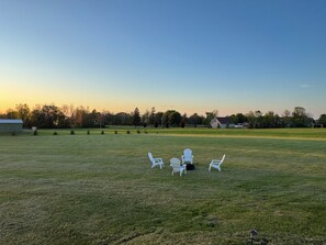 A grassy field with white chairs and a small table, surrounded by trees and houses.