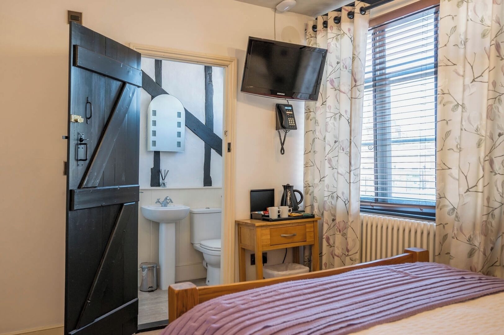 Un dormitorio con una cama, una mesita de noche con lámpara, un televisor montado en la pared, una ventana con cortinas y una puerta con diseño original.