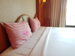 Une chambre d’hôtel avec un lit, deux oreillers roses, une tête de lit et une fenêtre avec des rideaux.