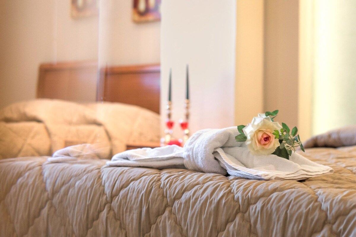 A neatly made bed with a white blanket, a folded towel, and a bouquet of flowers on the bed.