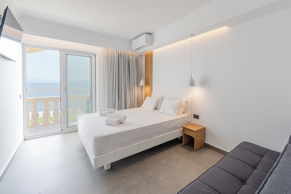 A modern bedroom with a large bed, a wooden nightstand, a grey sofa, and a balcony with curtains.