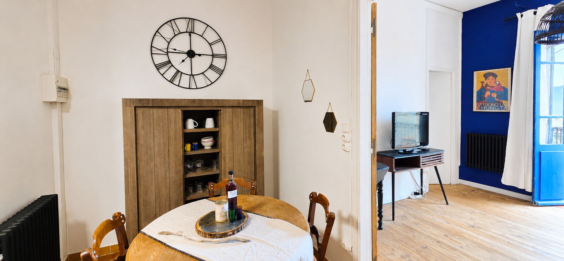 Een kamer met een eettafel, stoelen, een wandklok, een televisie en een blauwe accentmuur.