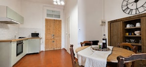 A compact kitchen with a microwave, oven, and wooden door. There's a dining table with chairs and a clock on the wall.