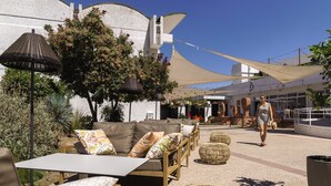 An outdoor seating area with sofas, tables, and wicker ottomans, shaded by large canopies and surrounded by plants.