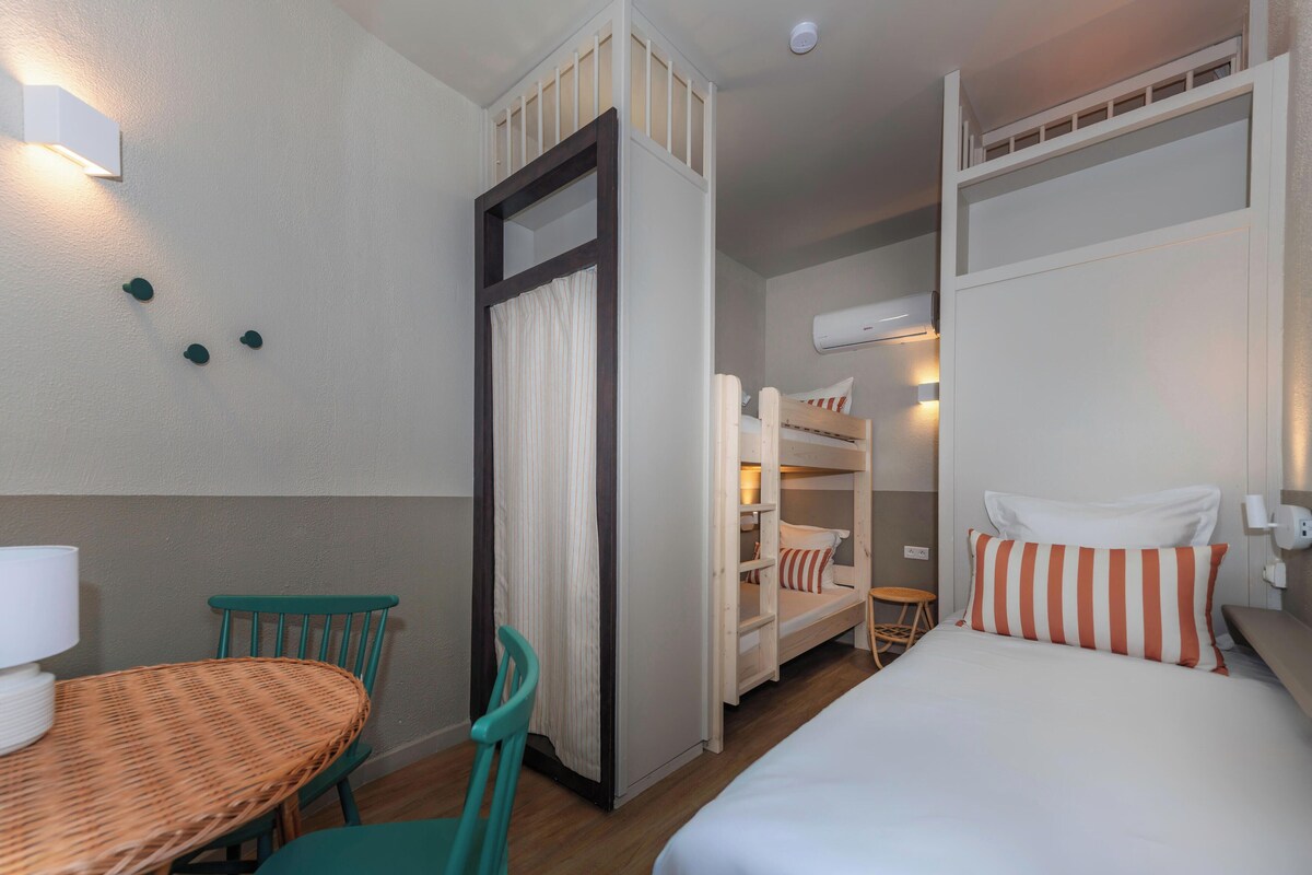 A hotel room with a bunk bed, a single bed, a dining table with chairs, and a small stool.