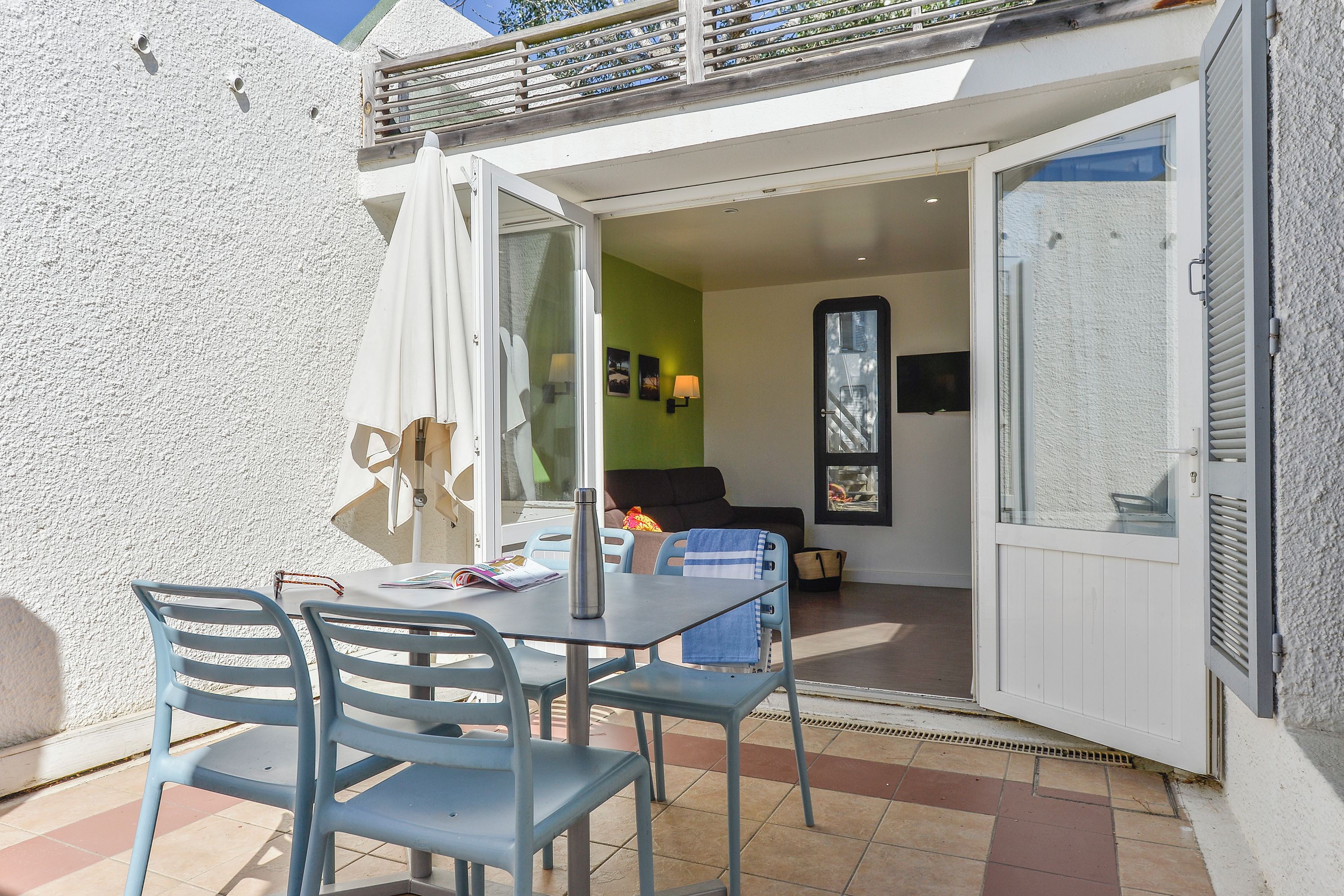 Une terrasse ouverte avec une table et des chaises, une porte-fenêtre coulissante donnant sur une pièce avec un canapé et un téléviseur, et un balcon avec une balustrade.