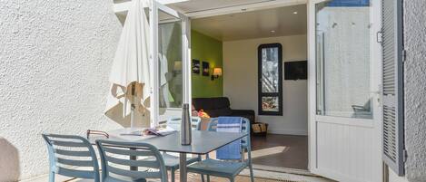 Une terrasse ouverte avec une table et des chaises, une porte-fenêtre coulissante donnant sur une pièce avec un canapé et un téléviseur, et un balcon avec une balustrade.