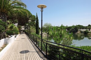 A wooden boardwalk with a metal railing, a lamp post, and a body of water with greenery.
