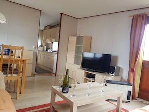 Ein Wohnzimmer mit Fernseher, einem Esstisch und einer Küche im Hintergrund.