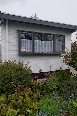 Ein Haus mit einem fensterförmigen Spalt, verziert mit einer Spitzenschürze, umgeben von unterschiedlichen Pflanzen und Blumen.