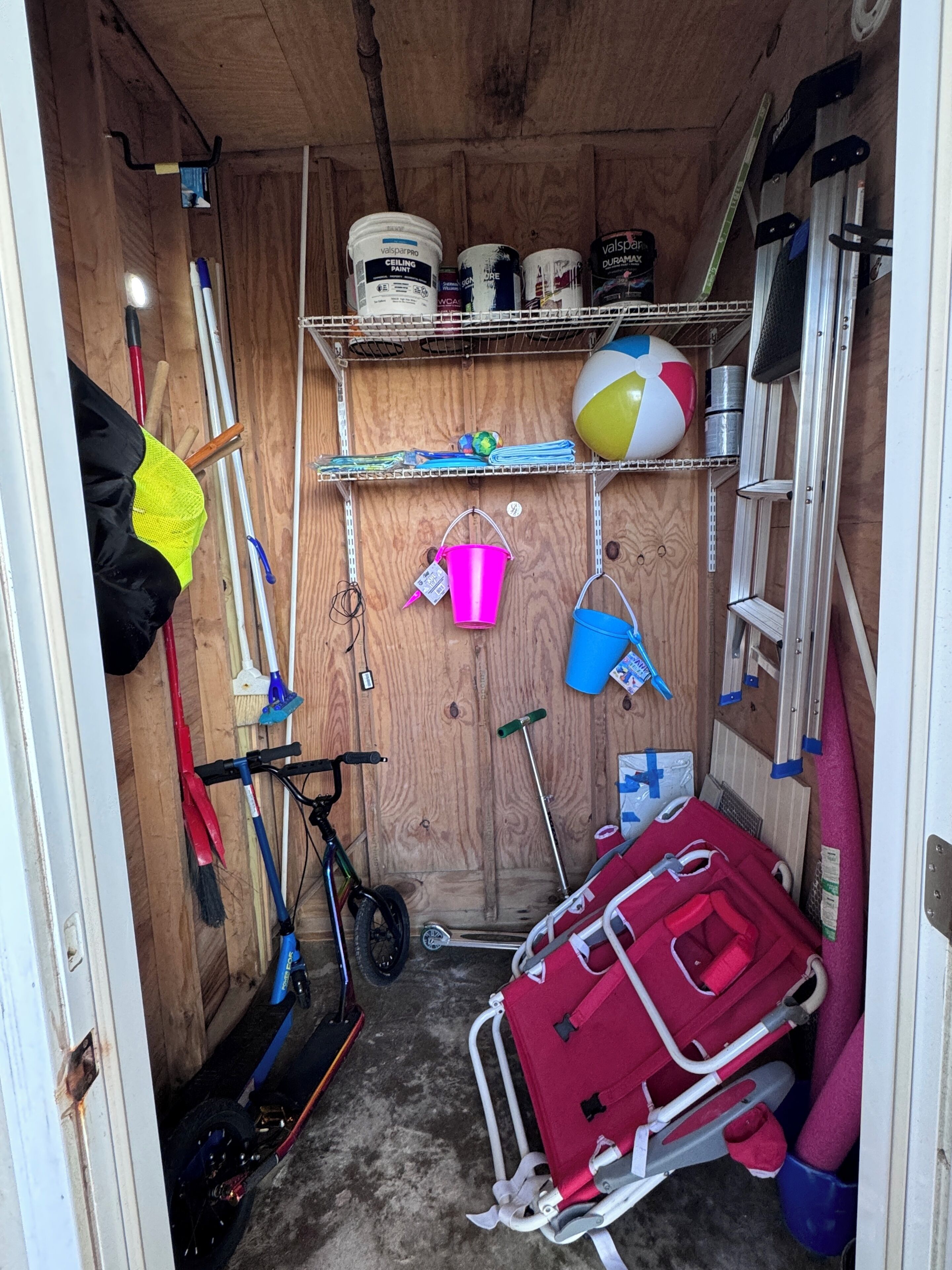 Un espace de rangement doté d’étagères où sont entreposés divers objets, comme des boîtes de peinture, un ballon de plage et des outils de nettoyage.