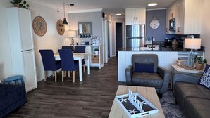 A modern living room with a wooden coffee table, a sofa, and a dining area with chairs and a table. - Welcome to Sea Clearly, a newly remodeled first floor oceanfront condo in OIB (Ocean Isle Beach)