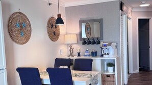 A dining area with a white table, blue chairs, a hanging lamp, and decorative wall hangings. - Welcome to Sea Clearly, a newly remodeled first floor oceanfront condo in OIB (Ocean Isle Beach)