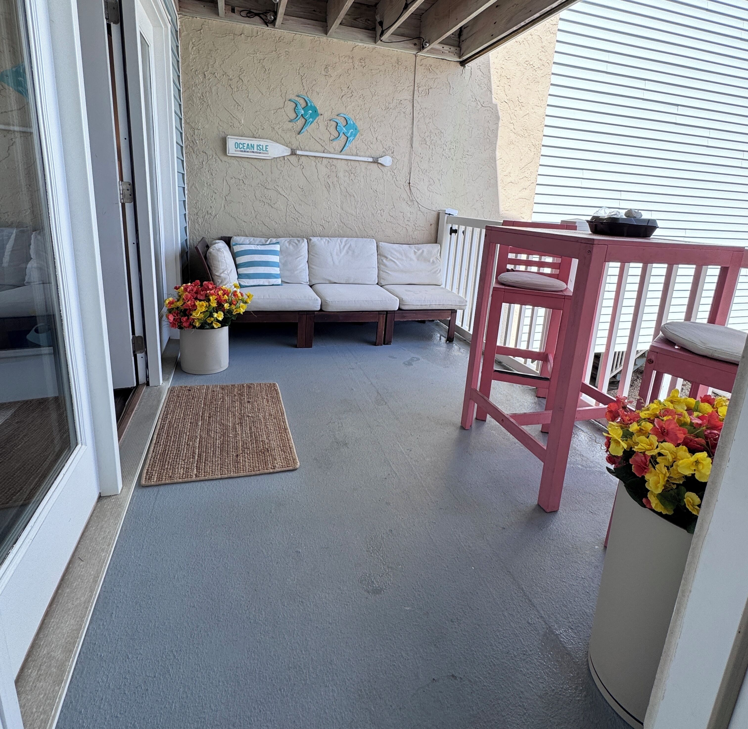 A covered outdoor seating area with a sofa, a chair, a doormat, and a wall-mounted sign.