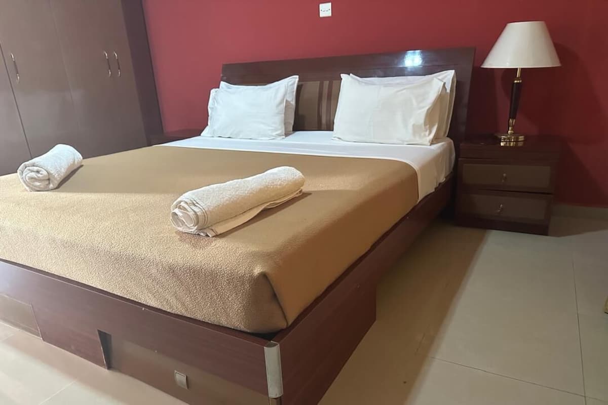 Ein ordentlich bezogenes Bett mit einer beigen Decke und weißen Kissen, ein Nachttisch aus Holz mit einer Lampe und eine rote Wand.