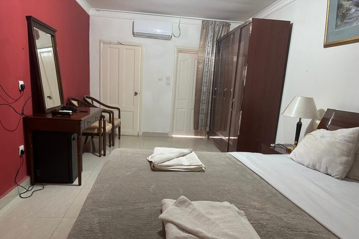 A hotel room with a bed, a desk with a mirror, a chair, a wardrobe, and a lamp.