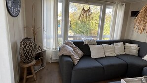 Ein Wohnzimmer mit einem dunkelgrauen Sofa, einer runden Wanduhr und Blick auf Bäume durch große Fenster.