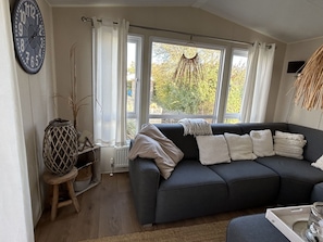 Ein Wohnzimmer mit einem dunkelgrauen Sofa, einer runden Wanduhr und Blick auf Bäume durch große Fenster.