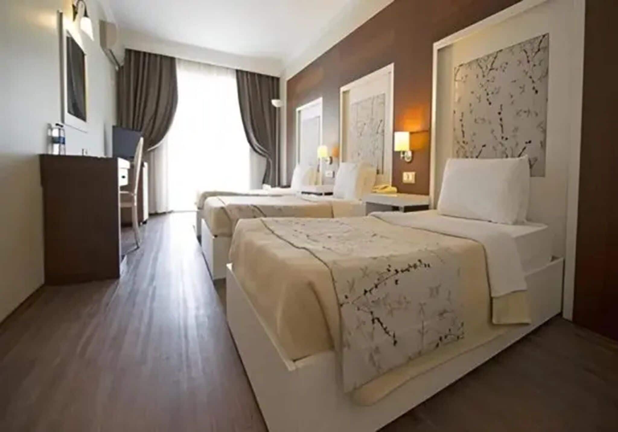 A hotel room with two beds, a desk, a chair, a wardrobe, and a large window with curtains.
