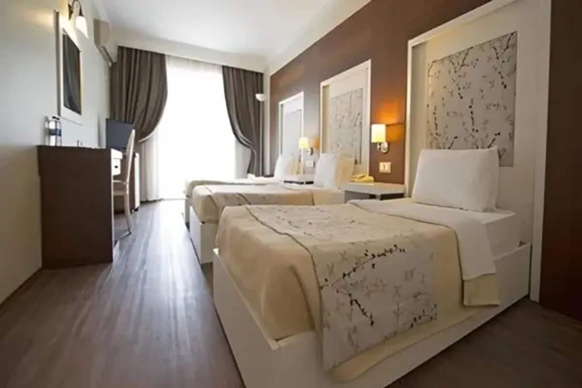A hotel room with two beds, a desk, a chair, a wardrobe, and a large window with curtains.