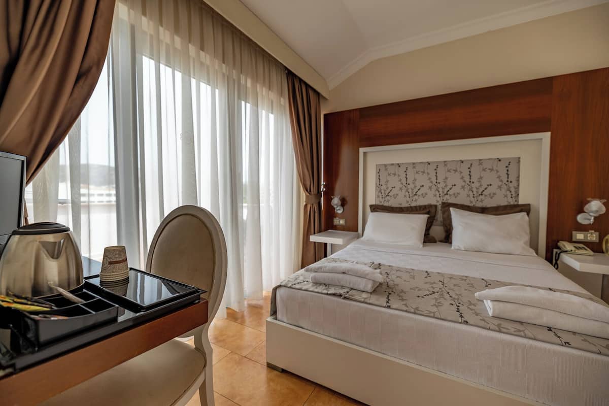 A hotel room with a bed, a desk with a kettle and cups, a chair, and a nightstand with a lamp.