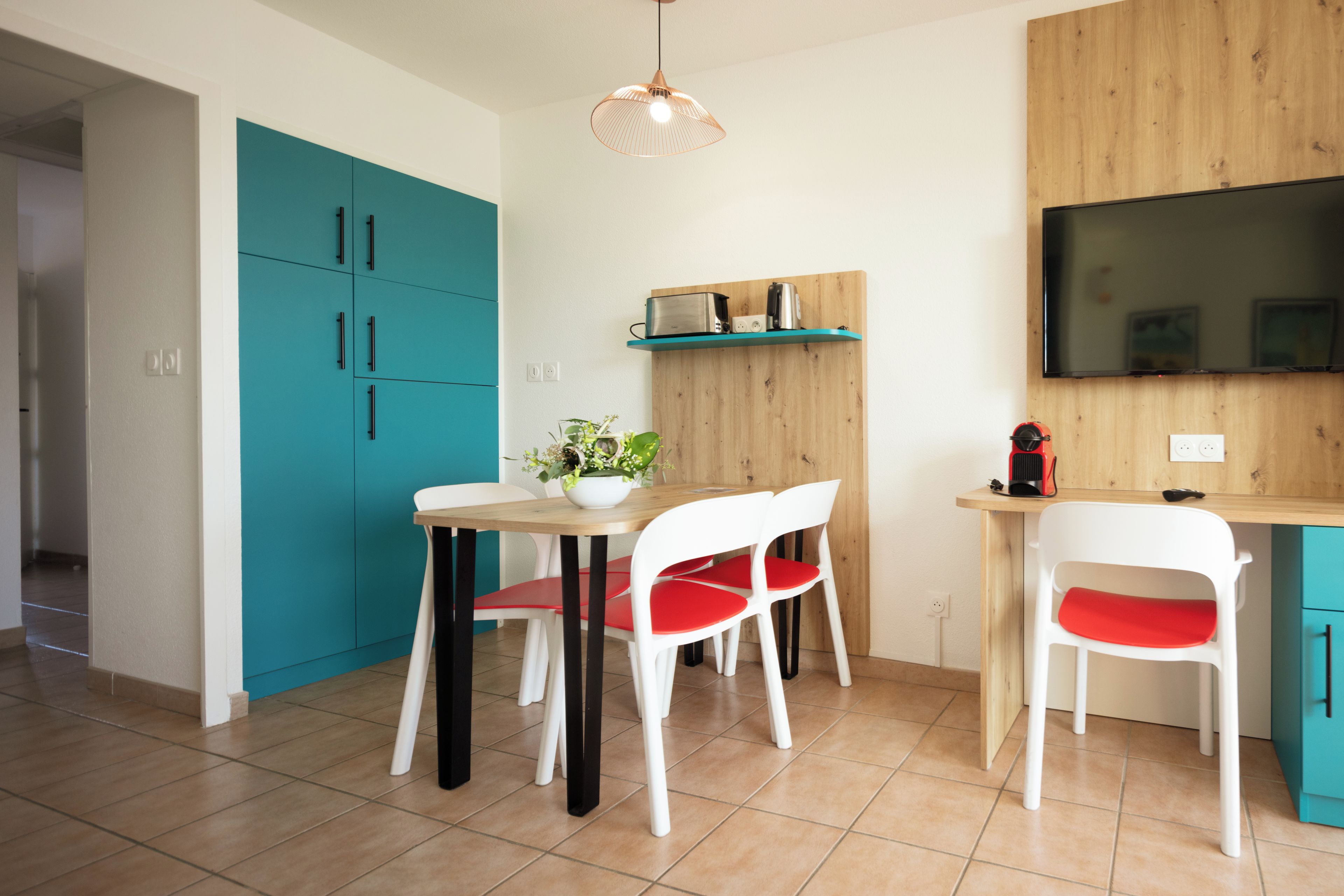 Una cocina moderna con armarios color turquesa, encimera de madera, una mesa de comedor con sillas blancas y un televisor montado en la pared.