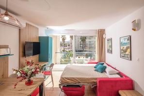 Ein Hotelzimmer mit Bett, Schreibtisch, Stuhl, Fernseher und einem Balkon mit Blick auf Palmen und Gebäude.