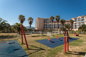 A playground with swings and slides, a fenced area, and a hotel building with balconies in the background. - Résidence Appart-hôtel Argelès-sur-mer (Argelès-sur-Mer)