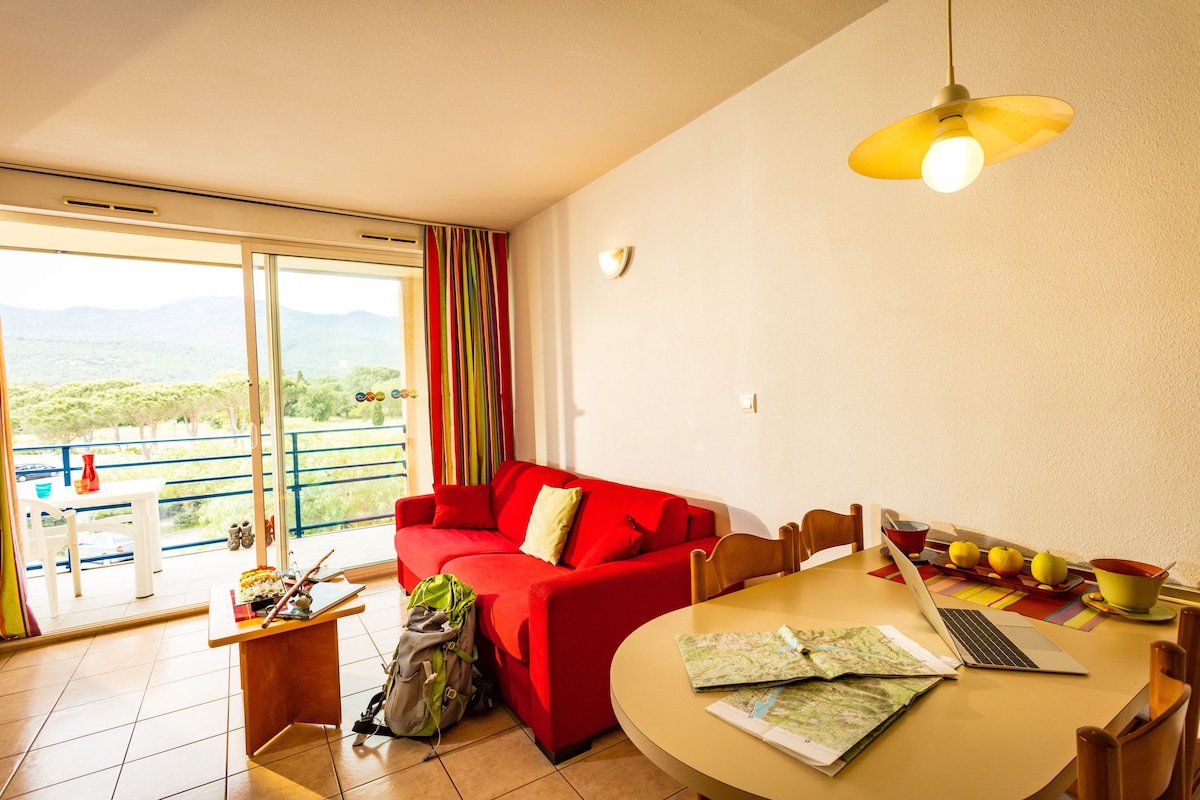 A room with a red sofa, a dining table with a laptop and fruits, and a view of greenery through large windows.