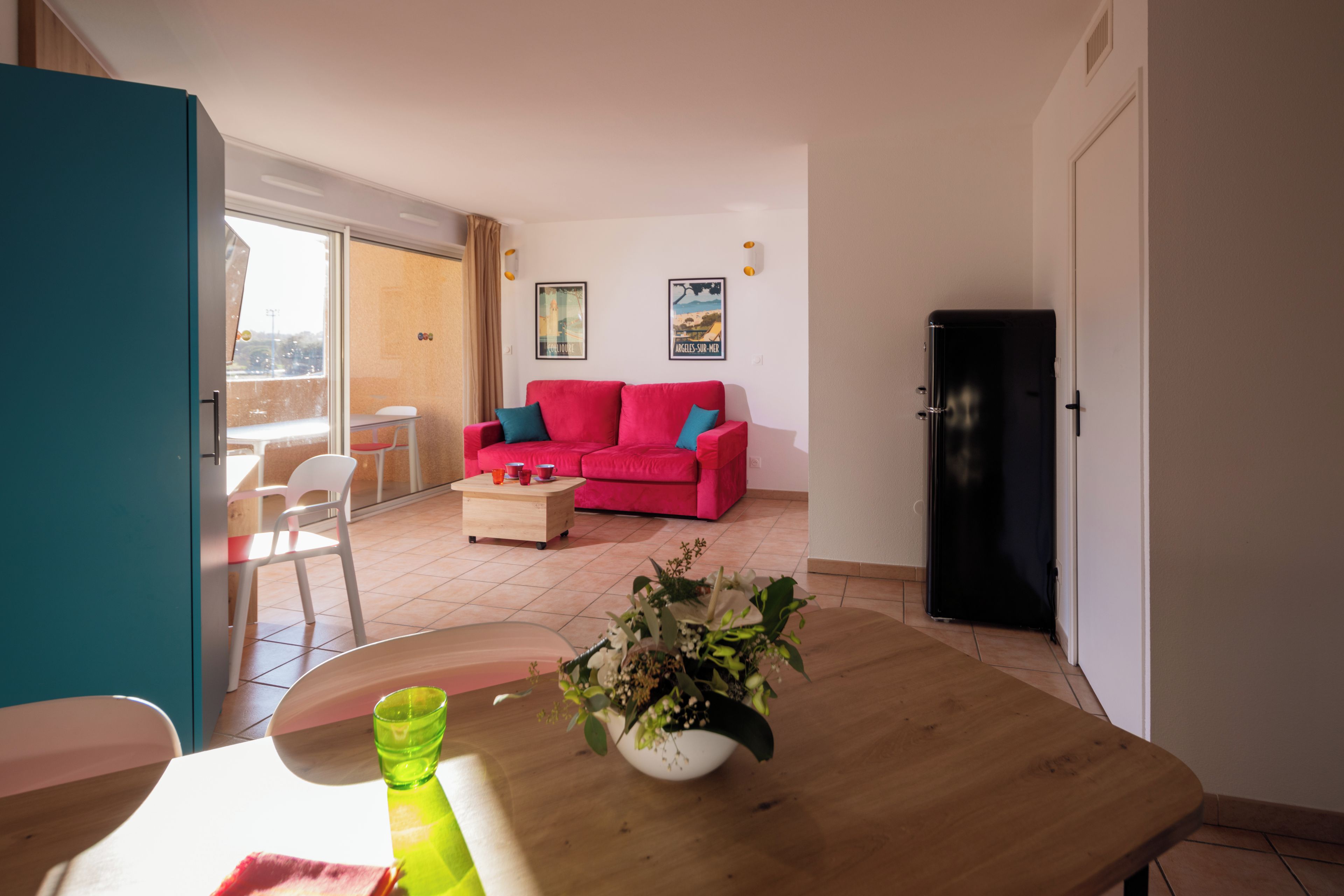 Una sala de estar con un sofá rojo, una mesa de comedor de madera y un florero con flores, además de un refrigerador negro.