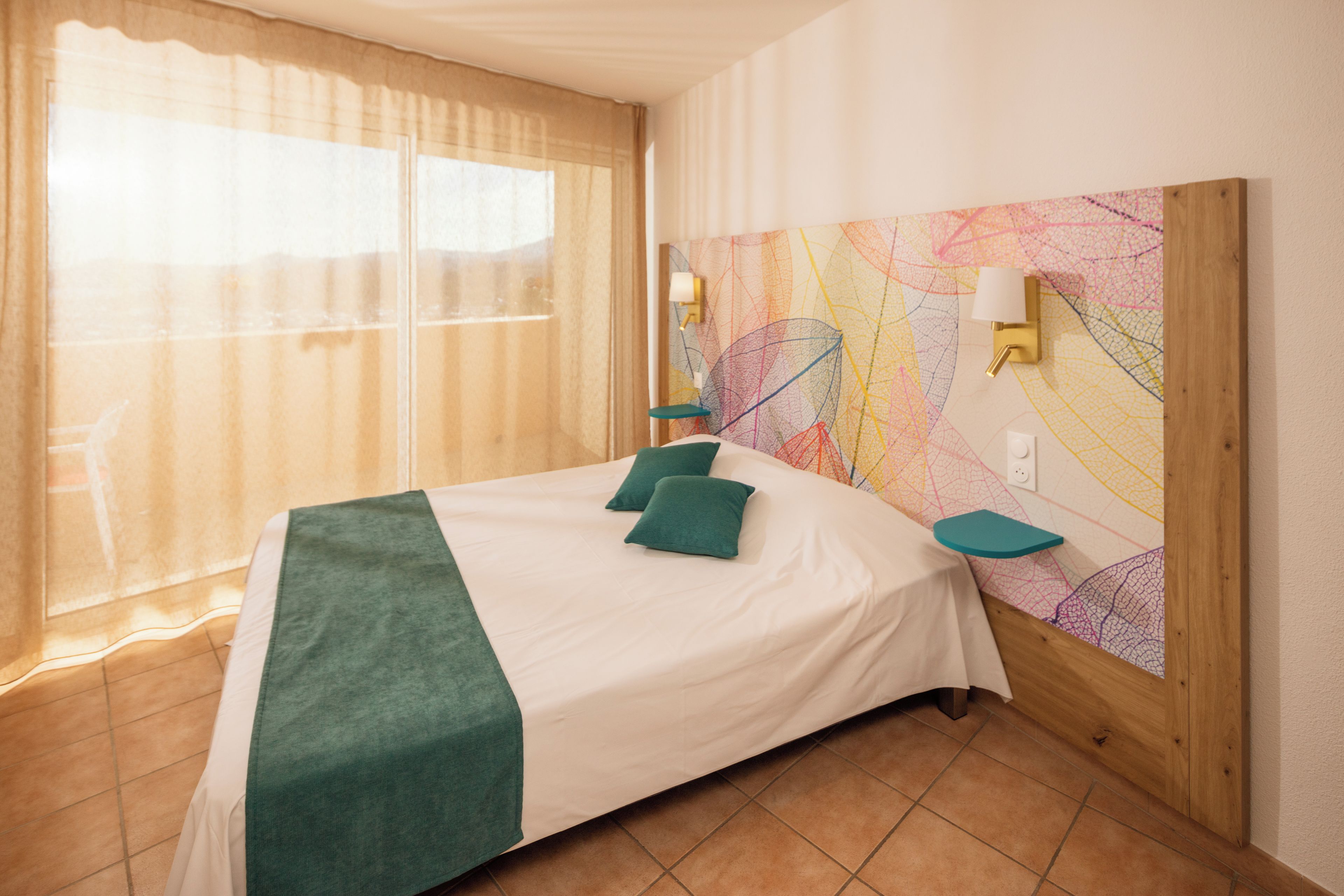 A hotel room with a bed, a window with curtains, a wall with a colorful abstract design, and a nightstand with a lamp.