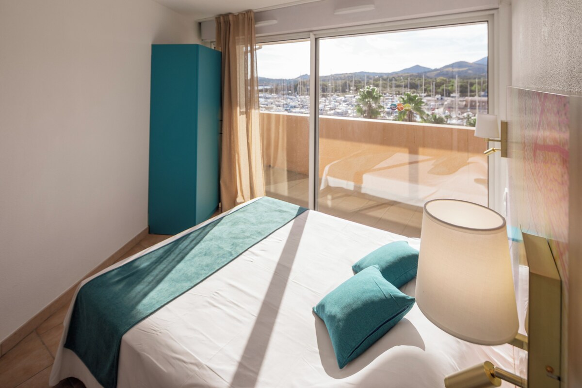 Habitación de hotel con cama, balcón con vista a un estacionamiento y montañas, y un ventanal con cortinas.