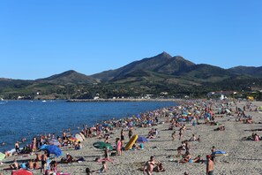 Een druk strand vol mensen die zonnebaden en zwemmen, met een berg op de achtergrond en een helderblauwe lucht.