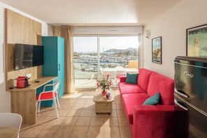 Una sala moderna con un sofá rojo, un televisor de pantalla plana, un refrigerador Cobalt, un escritorio de madera y vistas a un puerto deportivo a través de puertas de cristal grandes.