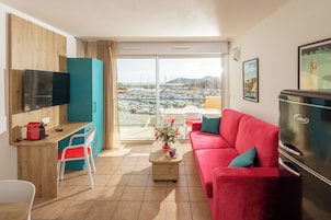 Un salon moderne avec un canapé rouge, un téléviseur à écran plat, un réfrigérateur de la marque Cobalt, un bureau en bois et une vue sur une marina, visible à travers de grandes portes vitrées.