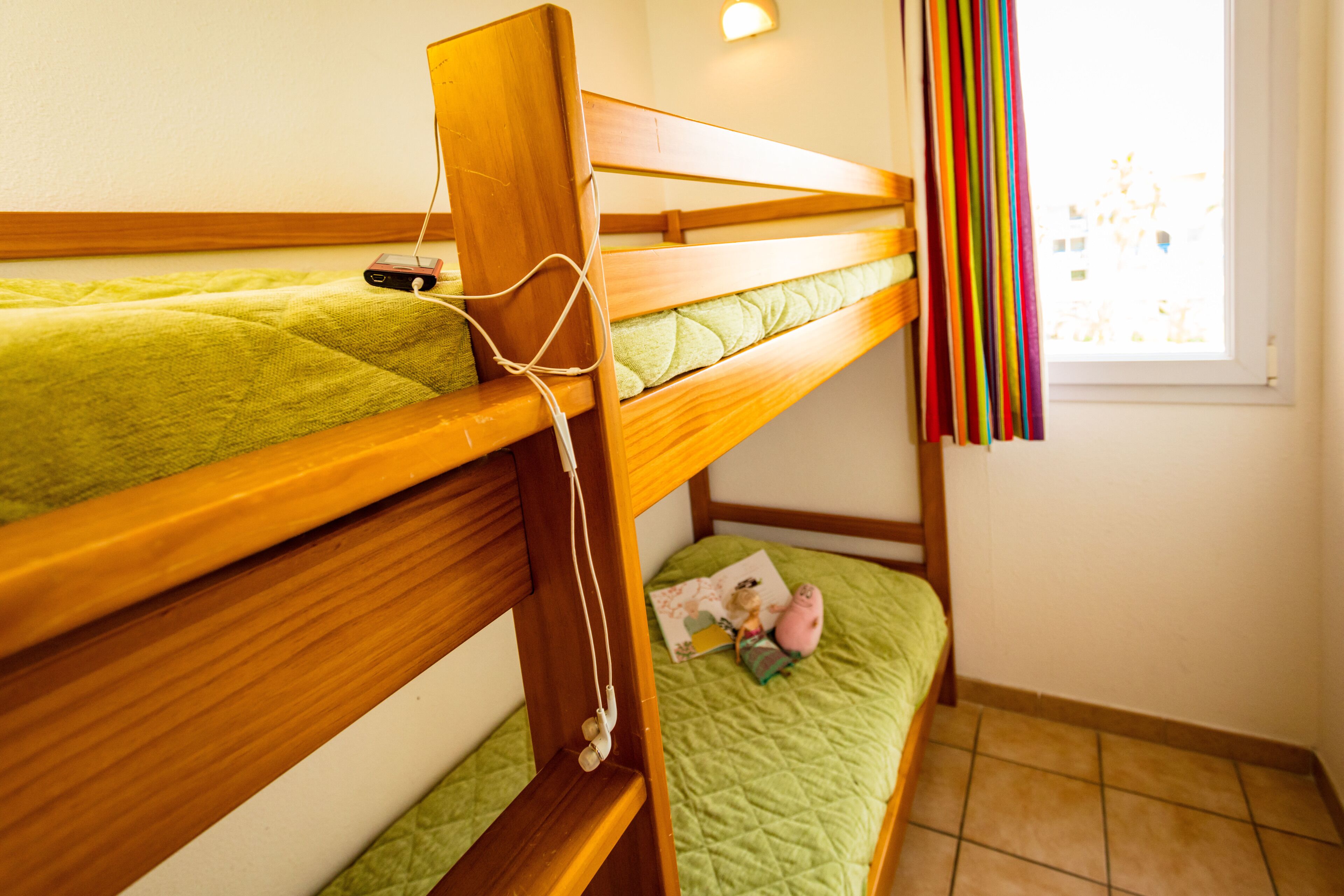 A bunk bed with a green quilt, a bedside table with a pink toy, and a window with colorful curtains.